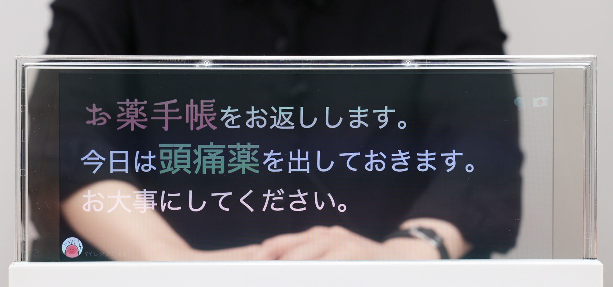 DNP対話支援システムの画面。透明な液晶ディスプレイに話した内容が表示され、「お薬手帳」や「頭痛薬」の文字は目立つフォントで表示されている。